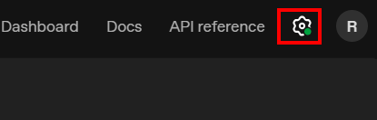 Captura de tela da interface escura do painel da OpenAI, mostrando a barra superior com os menus “Dashboard”, “Docs” e “API reference”. À direita, um ícone de engrenagem com um ponto verde está destacado por um retângulo vermelho, próximo ao avatar circular com a letra R