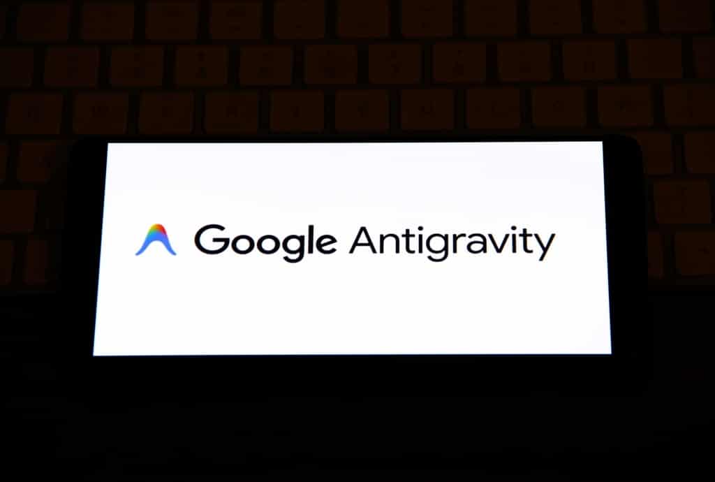 Fotografia em close de um smartphone posicionado horizontalmente sobre um teclado de computador em um ambiente com baixa luminosidade. A tela do aparelho está acesa e exibe o logotipo colorido e o nome Google Antigravity em destaque sobre um fundo branco brilhante. A composição sugere o uso da ferramenta em dispositivos móveis e a integração com o ecossistema de trabalho do desenvolvedor.