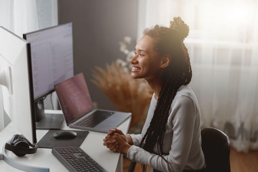 Uma mulher jovem com cabelos trançados sorri enquanto trabalha em seu escritório doméstico bem iluminado. Ela está sentada à frente de um monitor grande e um laptop, ambos exibindo linhas de código e estruturas de automação. O ambiente sugere o processo de estudo e implementação técnica necessário para customizar templates n8n de acordo com as necessidades de integração de dados e sistemas.