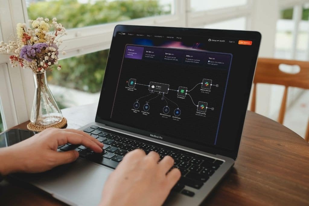 Visão aproximada de mãos digitando em um laptop que mostra a montagem de um fluxo complexo utilizando templates n8n. No centro da tela, destaca-se um nó de AI Agent conectado a diversas ferramentas como Microsoft Excel, Jira e Slack. A interface demonstra visualmente como as automações de inteligência artificial podem ser estruturadas para enviar atualizações de perfil e mensagens em canais de comunicação.